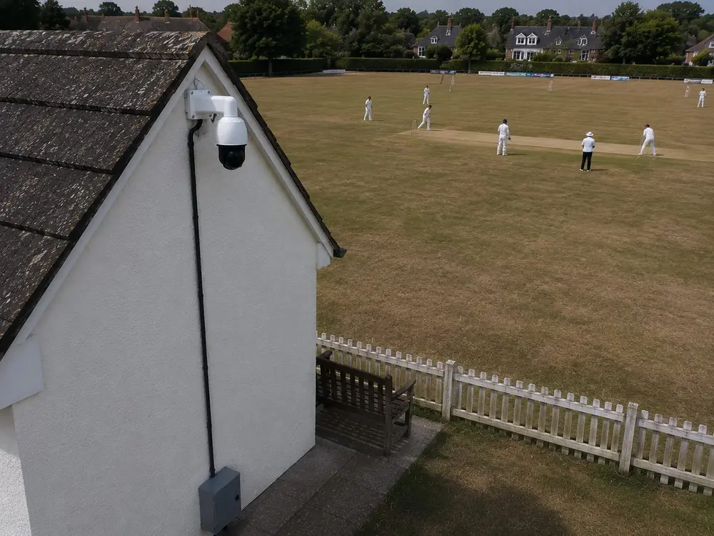 Streaming camera mounted on a pavilion gable end with the cable run down to a sealed equipment cabinet, cricket match in play beyond